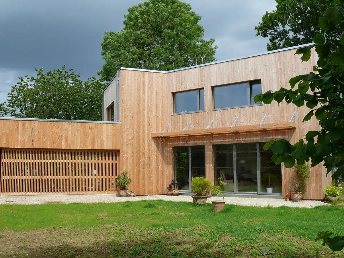 Une maison passive moderne en bois, avec de grandes baies vitrées et une porte de garage ajourée, entourée de verdure.