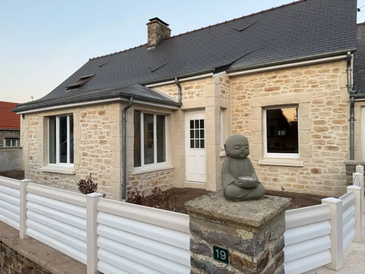 Façade maison traditionnelle en pierre avec une statue de Bouddha méditant. Une clôture blanche orne l'entrée.