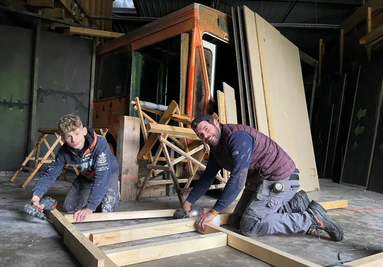 Deux hommes travaillent sur un cadre en bois, un projet de recyclage du bois de construction. Derri&egrave;re eux, un vieux camion rouill&eacute; et des panneaux de bois.