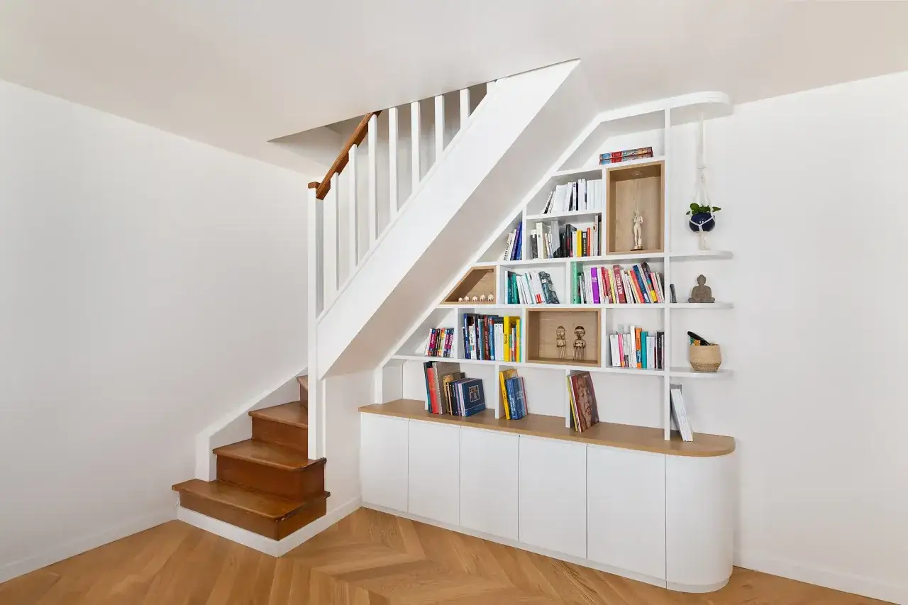 Bibliothèque intégrée sous un escalier, remplie de livres et d'objets décoratifs. Des portes blanches ferment la partie basse.