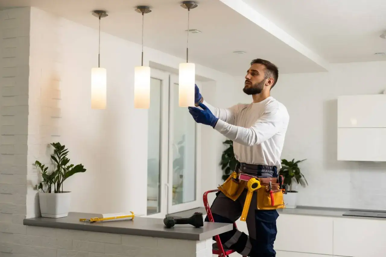 Un &eacute;lectricien artisan installe des luminaires modernes. Il est sur un escabeau, portant des gants bleus et une ceinture &agrave; outils.
