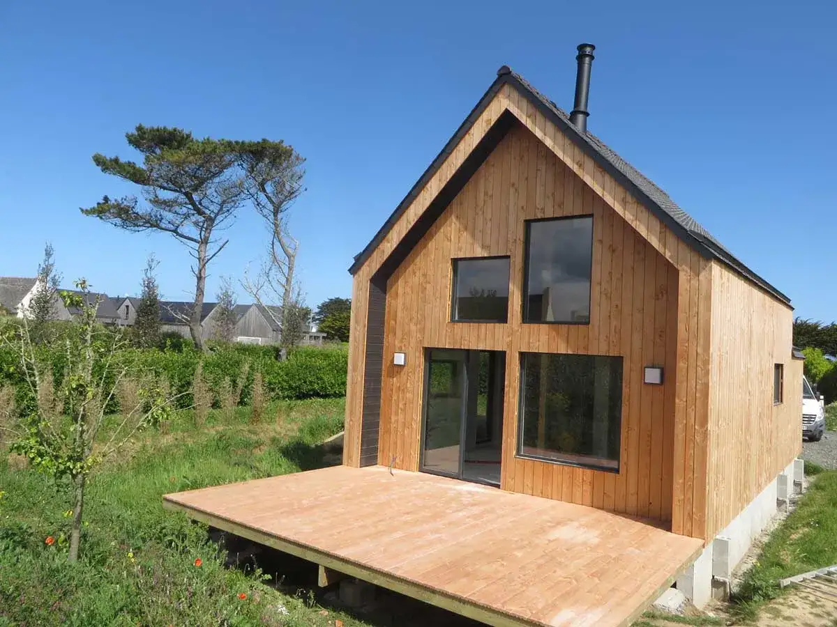 Une petite maison moderne en bois avec une terrasse, entourée de verdure et sous un ciel bleu.