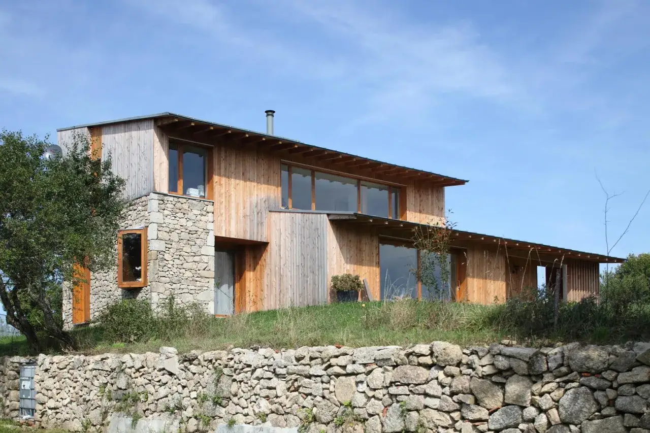 Maison de campagne simple en bois et pierre, avec de grandes fenêtres et un toit débordant, nichée dans un paysage verdoyant.