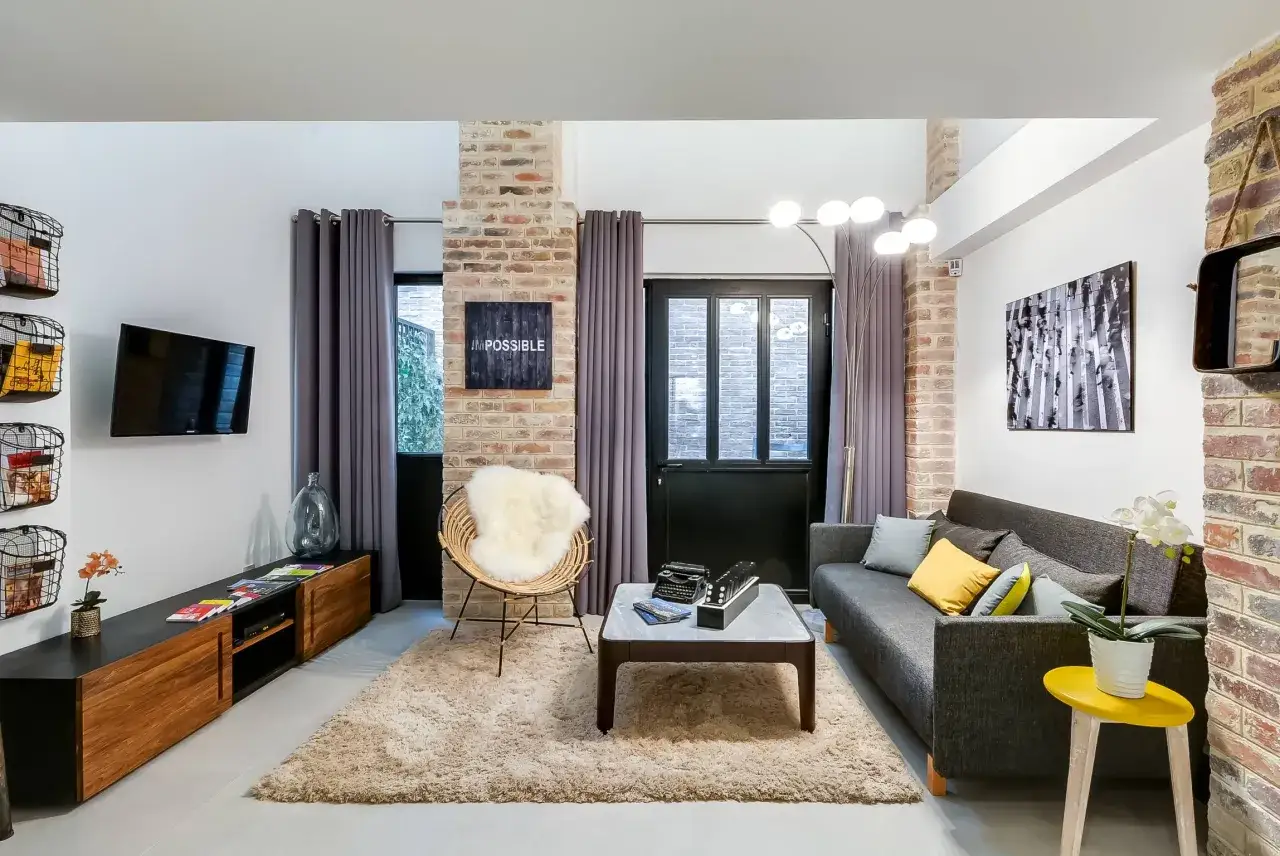 Salon moderne avec murs en briques apparentes. Un canap&eacute; gris, un fauteuil en rotin, une table basse avec une machine &agrave; &eacute;crire et des dominos. 1 ou 2 rideaux par fen&ecirc;tre.