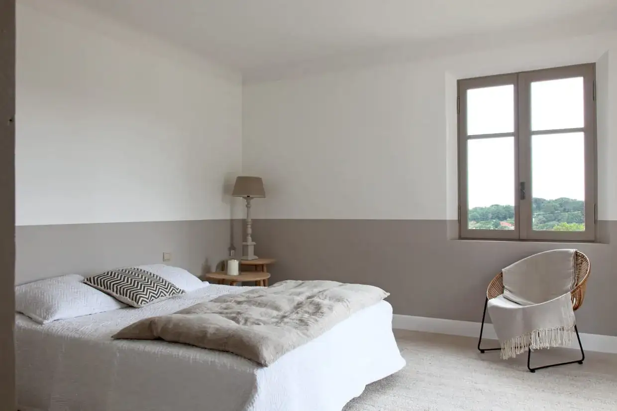 Chambre paisible avec un lit blanc, un fauteuil en rotin et un mur bicolore. Le soubassement du mur est peint en gris taupe, contrastant avec le blanc du haut.