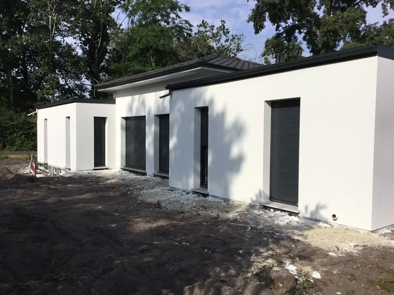 Maison moderne avec toit plat, murs blancs et volets roulants gris. Le terrain autour est en cours d'aménagement.