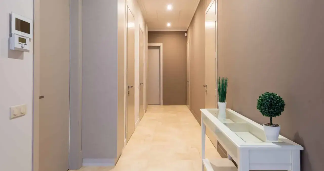 Un couloir aux murs peints en beige et gris, avec plusieurs portes. Un meuble blanc orné de plantes vertes se trouve sur la droite.