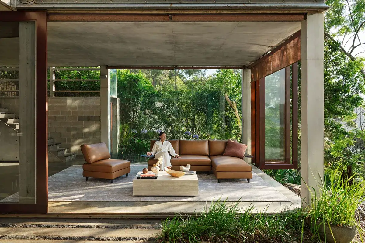 Un salon ouvert sur la nature, avec un canap&eacute; en cuir marron et une femme assise. Ce logement du futur s'int&egrave;gre parfaitement &agrave; son environnement verdoyant.