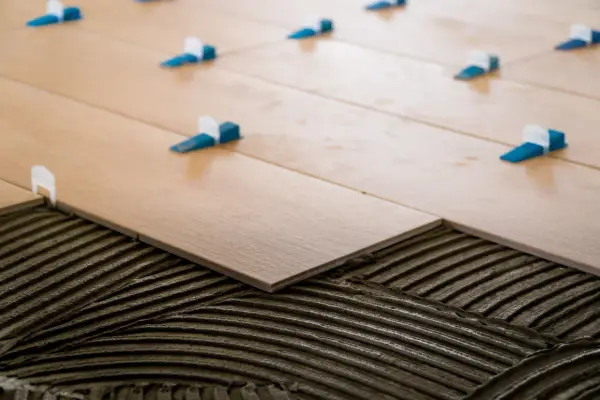 Pose de carrelage 60x60 en cours. Des carreaux en bois clair sont posés sur du mortier frais, avec des croisillons bleus et blancs pour un espacement parfait.