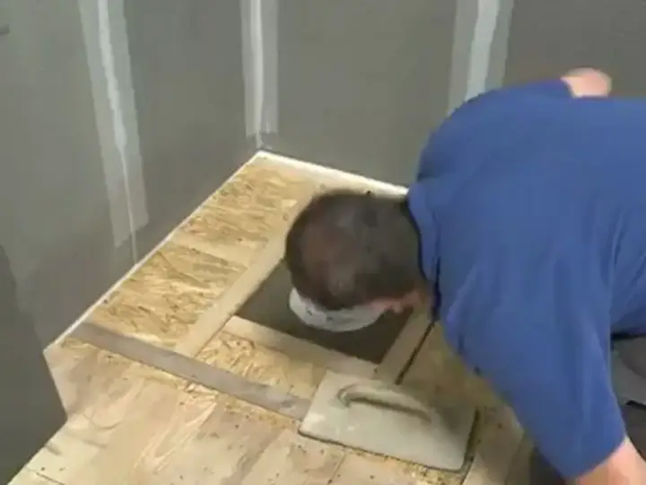 Un homme installe une douche sur un plancher en bois brut. Il utilise une truelle pour étaler du mortier autour du drain.