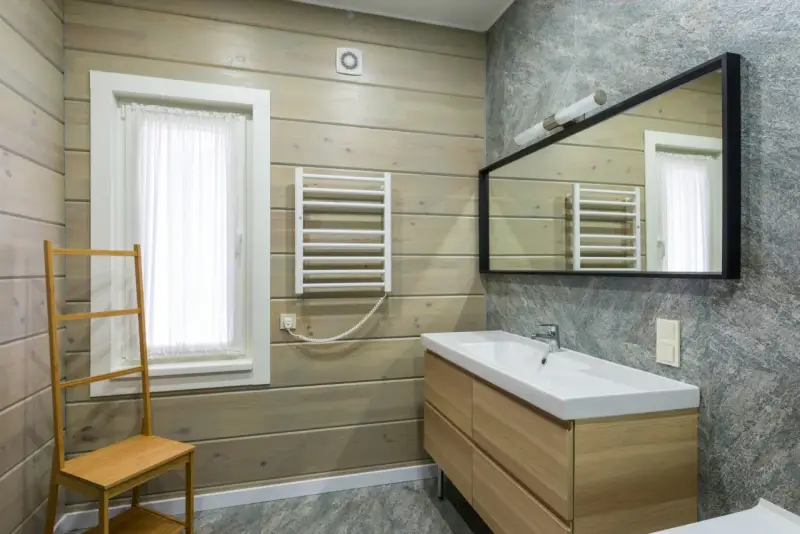 Salle de bain moderne avec murs en bois clair et pierre grise. Un meuble vasque en bois, un miroir, un radiateur et une échelle en bambou pour habiller un mur.