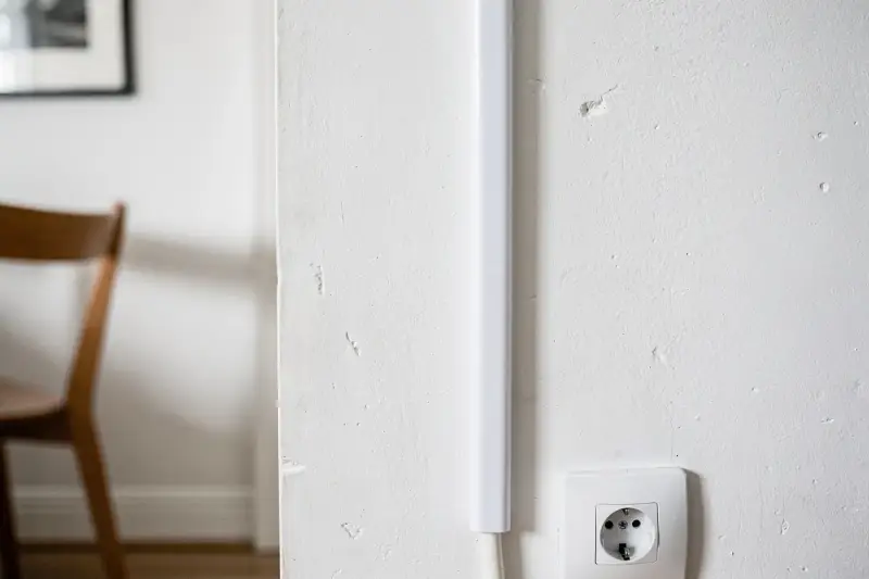 Une goulotte blanche est fixée verticalement sur un mur blanc, dissimulant des câbles. Une prise électrique est visible en bas à droite.